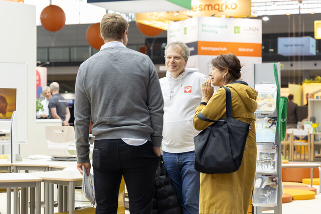 Ein Fachberater von ASS Einrichtungssysteme unterhält sich auf der Didacta neben einer Tischgruppe mit zwei Messebesuchern