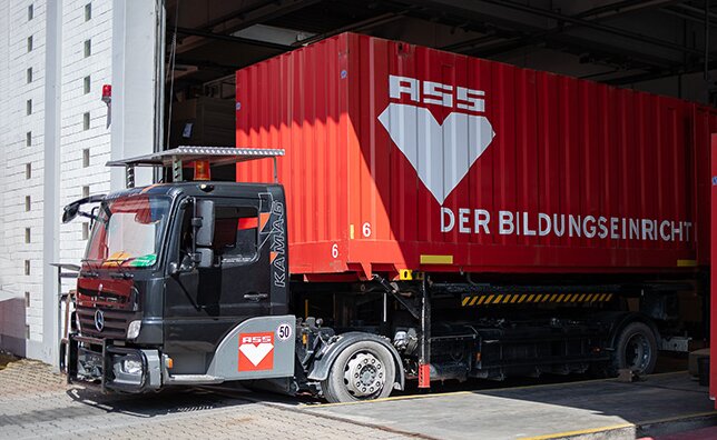 Anfahrender LKW mit rotem Container und ASS-Einrichtungssysteme GmbH-Logo