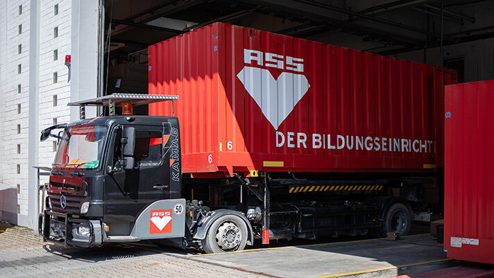 Anfahrender LKW mit rotem Container und ASS-Einrichtungssysteme GmbH-Logo